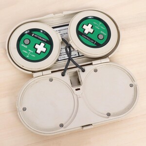 May include: An open, off-white plastic device with two circular controllers. Each controller has a green center with a white cross-shaped button and directional labels. The text "SPRAY" and "CONTROLLER" are visible. A gray cord connects the two controllers.