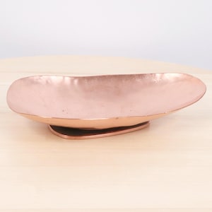 May include: An oval-shaped copper bowl with a smooth, metallic finish and a slightly irregular, organic rim. The bowl features a shallow, wide basin and rests on a small, integrated base, showcasing a warm, reflective rose-gold colour throughout.