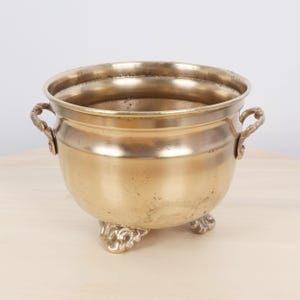 Peut inclure: Un pot en métal de couleur laiton avec un corps arrondi, deux poignées ornées et trois pieds décoratifs. Le pot a une large ouverture et une finition légèrement ternie, suggérant l'âge. Style vintage ou antique.