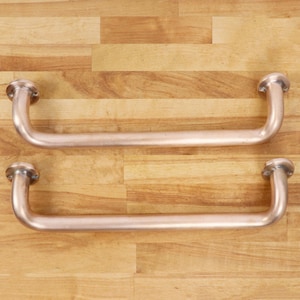 Può includere: Due maniglie in metallo curve color rame con piastre di montaggio circolari. Le maniglie sono posizionate orizzontalmente su una superficie di legno marrone chiaro. Il design è semplice e industriale.