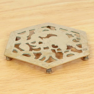 May include: A hexagonal brass coaster with a decorative floral design. The coaster has six legs and is made of a shiny metal.