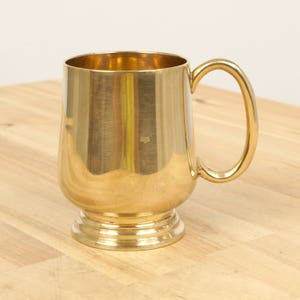 Puede incluir: Una taza de metal de color dorado con un cuerpo redondeado y un asa. La taza tiene un acabado liso y pulido.