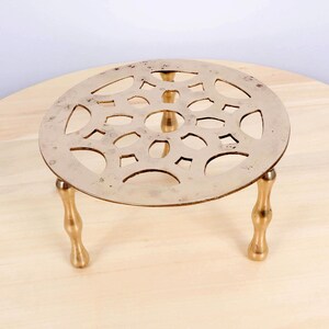 May include: A circular brass trivet with an openwork pattern, supported by three short, curved legs. This heat-resistant trivet, designed to protect surfaces, is shown on a light wooden surface. The trivet's diameter is approximately 15 cm.