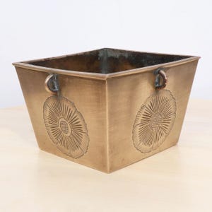 May include: A square, bronze-coloured metal planter with a slightly tapered design. The planter features two decorative floral designs etched on the front and side panels. Small, curved handles are attached to the top rim.