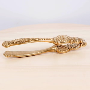 May include: A gold-colored metal nutcracker featuring an ornate, embossed floral pattern along the handles and a textured grip area. The tool has a hinged design with a central section containing vertical ridges for cracking shells.