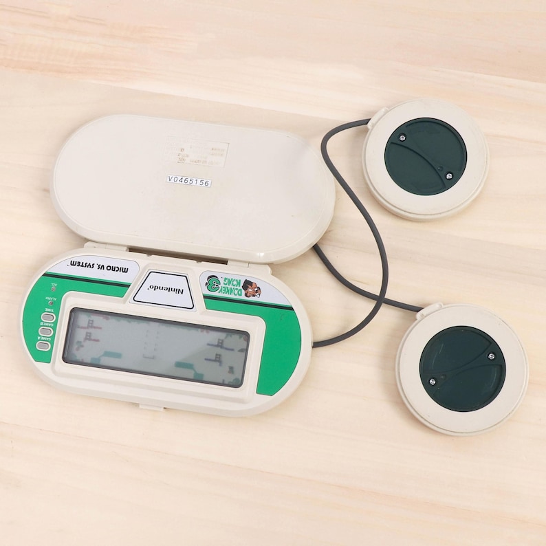 May include: A vintage Nintendo Donkey Kong Micro Vs. System handheld game. The game is white and green with a digital display showing the game's graphics. Two circular controllers are connected to the main unit by a gray cable.