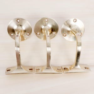 May include: Three brass-colored curtain rod holders. Each holder has a circular mounting plate with screw holes, a curved arm, and a rectangular base with screw holes. The holders are arranged on a light-colored wooden surface.