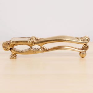 May include: A gold-coloured nutcracker with ornate, scrolled detailing. The nutcracker has a lever design with a rectangular base and a rounded end. The overall design is vintage and decorative, suitable for kitchen or bar decor.