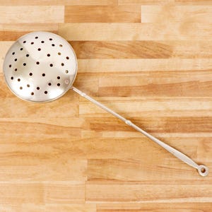 May include: A silver-colored metal skimmer spoon featuring a large, circular, perforated bowl attached to a long, slender handle. The handle terminates in a flat, circular loop at the end, designed for hanging the utensil when not in use.