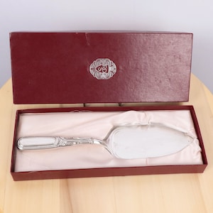 Puede incluir: Un cuchillo para pastel de plata con un mango adornado, presentado en una caja forrada de satén blanco. La caja tiene una tapa roja con un escudo dorado que dice "R.S."