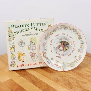 May include: A white ceramic Christmas plate with a red rim featuring Peter Rabbit and a festive scene. The plate has a Beatrix Potter inscription and the year 1983. The plate is displayed on a gold stand.