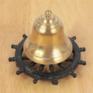 May include: A gold-colored bell with a black ship's wheel base. The bell is round and has a smooth surface. The base is made of metal and has a black finish.