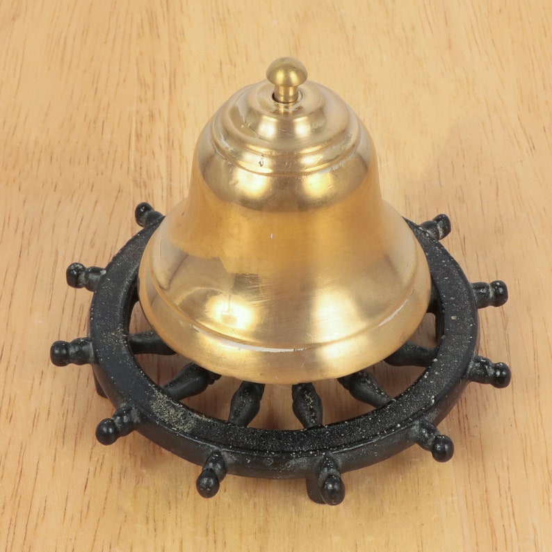 May include: A gold-colored bell with a black ship's wheel base. The bell is round and has a smooth surface. The base is made of metal and has a black finish.