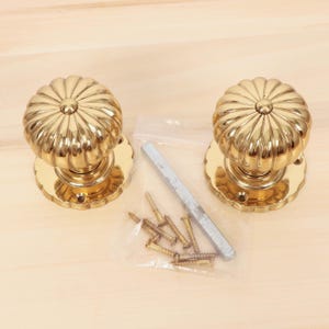 Op de afbeelding: Twee gouden deurknoppen met een geribbeld ontwerp. De knoppen bevinden zich op een houten oppervlak met een plastic zak die schroeven en een metalen staaf bevat.