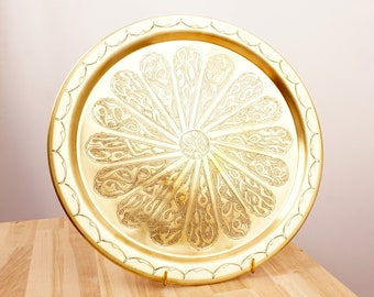 May include: A gold metal serving tray with an intricate, circular design. The design features a central flower with radiating petals, each with a detailed pattern.