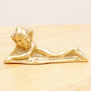 Peut inclure: Une figurine en métal doré représentant une personne allongée avec un visage souriant. La figurine est posée sur une surface en bois.