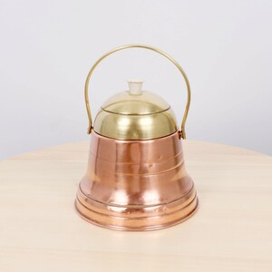 May include: A copper and brass ice bucket with a handle. The ice bucket has a copper base and a brass lid and handle. The handle arches over the top. The ice bucket is sitting on a light wood surface.