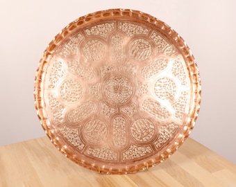 May include: A round copper serving tray with intricate engraved designs. The tray has a raised rim and a hammered finish.