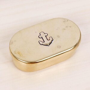 May include: An oval-shaped brass box with a polished metallic finish. The lid features a centered, engraved anchor symbol with dark detailing. The container has smooth, rounded edges and a solid, reflective surface throughout its exterior.