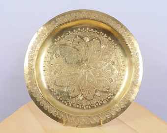 May include: A round, gold-colored metal tray with an intricate floral pattern etched into the surface. The tray has a decorative border and is displayed on a wooden surface. The tray is supported by two small gold-colored stands.