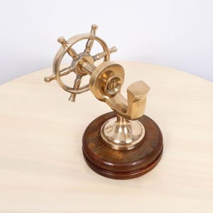 May include: A brass ship's wheel-shaped hook mounted on a round, dark wood base. The hook is designed to hold items and has a nautical theme. The brass has a polished finish, and the wood base is stained.