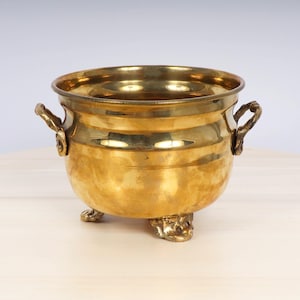 May include: A gold-toned brass planter with a rounded body and four decorative feet. The planter has two handles on the sides.