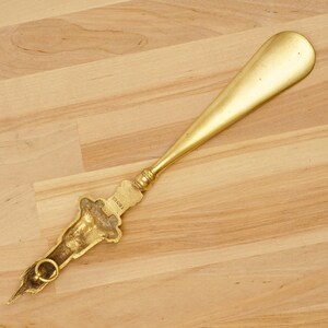 Peut inclure: Un chausse-pied en or avec une poign&eacute;e d&eacute;corative. La poign&eacute;e est en forme de personnage stylis&eacute; avec l'inscription "1889" dessus.