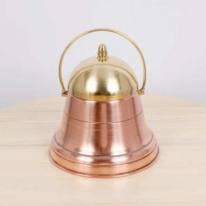 Peut inclure: Un récipient en forme de cloche en cuivre et laiton avec une poignée. Le dessus est de couleur dorée et le corps est en cuivre avec des bandes horizontales. La poignée est un arc de couleur dorée. Le récipient est posé sur une surface en bois de couleur claire.