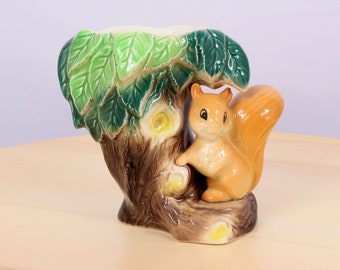 May include: A ceramic squirrel figurine perched on a tree trunk with green leaves. The squirrel is brown and orange with a bushy tail. The tree trunk is brown and has yellow flowers.