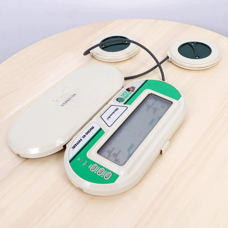 May include: A vintage Nintendo Micro VS System game console. The handheld device is white and green with a screen and buttons. Two circular controllers are connected by a black wire. The game is on a light wood surface.