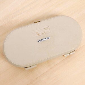 May include: A beige, oval-shaped plastic case with a hinged lid and a small rectangular label with the text "V0465156". The case has a smooth surface and a slightly raised edge. The case is closed.