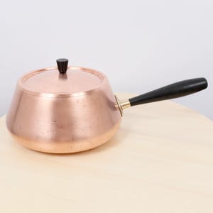 May include: A copper saucepan with a lid and a black wooden handle. The handle is attached with a brass fitting. The pan has a rounded body and a small, black knob on the lid.