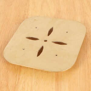 May include: A gold-colored metal square plate with a cut-out design in the center. The design is a four-pointed star with rounded edges.