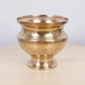 Op de afbeelding: Een goudkleurige metalen kom met een brede rand en een voetstuk. De body van de kom heeft een geëtst bloemmotief. De kom staat op een lichtgekleurd houten oppervlak.