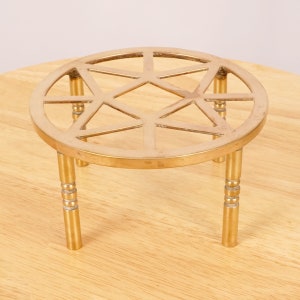 Puede incluir: Un soporte de metal dorado con una parte superior redonda y tres patas. La parte superior tiene un diseño geométrico con una forma de estrella en el centro.