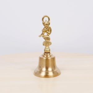 May include: A vintage brass bell with a mermaid figurine on top. The bell has a flared base and a loop for hanging. The mermaid is seated and holding her knees. The bell is a warm gold color and sits on a light-colored surface.