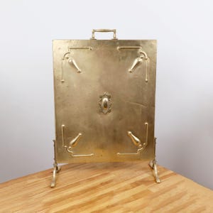 May include: A brass fireplace screen with a rectangular design. The screen has a handle at the top and decorative accents. It is supported by two legs and is placed on a wooden surface.