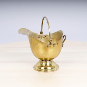 Könnte beinhalten: Ein kleiner, goldfarbener Metalleimer mit Henkel. Der Eimer hat einen abgerundeten Boden und einen ausgestellten Rand. Er steht auf einer hellbraunen Holzoberfläche.