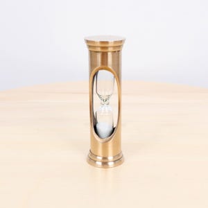 Puede incluir: Un reloj de arena de latón con un diseño cilíndrico y un recorte ovalado que revela la cámara de vidrio. El reloj de arena contiene arena blanca y está sobre una superficie de madera clara.