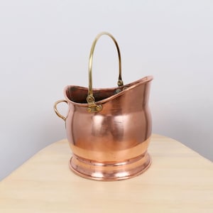 Puede incluir: Un cubeta de carbón de cobre con un asa de latón y un asa pequeña de latón en el lateral. La cubeta tiene un cuerpo redondeado y una base acampanada. El cobre tiene una superficie brillante y reflectante.