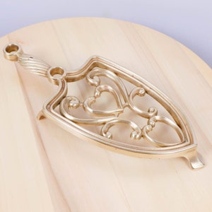 Puede incluir: Un salvamanteles de metal dorado con un diseño decorativo de corazón. El salvamanteles tiene un asa en un extremo y está diseñado para proteger las superficies de los platos calientes.