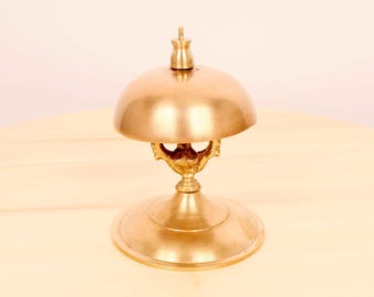 Op de afbeelding: Een gouden messing hotel-servicebel met een ronde bovenkant en een decoratieve basis. De bel staat op een licht houten oppervlak.