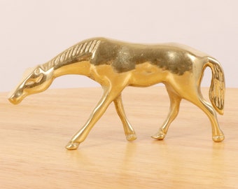 May include: A solid brass horse figurine with a detailed mane and tail. The horse is depicted in a walking pose with its head lowered.