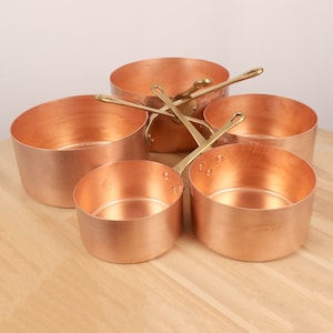 May include: Set of four copper saucepans with brass handles. The saucepans are nested together and have a shiny, polished finish.