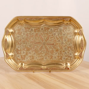 May include: A gold-coloured metal serving tray with an ornate floral design. The tray has a raised edge and a slightly curved shape.