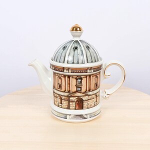 Op de afbeelding: Een witte keramische theepot met een gedetailleerde illustratie van een gebouw met een koepel. De theepot heeft een goudkleurige knop en gouden rand op het handvat. De gebouwillustratie gebruikt bruine, beige en blauwe tinten.