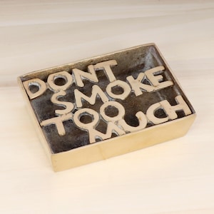 Op de afbeelding: Een gouden metalen asbak met de woorden "DON'T SMOKE TOO MUCH" in reliëf op de bovenkant.