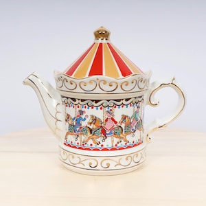 Op de afbeelding: Een witte theepot van keramiek met een rood en geel carrouselontwerp. De theepot heeft een gouden rand en een handvat. De carrousel toont kinderen op paarden.