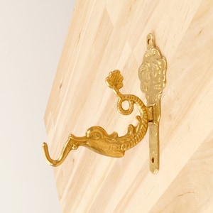 May include: A gold-colored, ornate wall hook featuring a seahorse design. The hook is mounted on a wooden surface. The seahorse has a curved tail and a detailed head. The hook is designed for hanging items.
