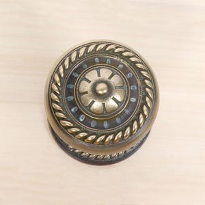 Peut inclure: Un bouton de meuble rond en laiton orné d'un motif floral central entouré d'une bordure en forme de corde. La surface métallique présente une patine sombre dans les zones en creux, contrastant avec les reflets polis des détails en relief.